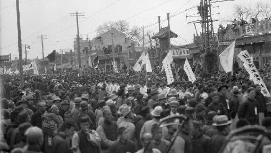 A black and white image shows a street full of Chinese crowds with other people watching on on rooftops.