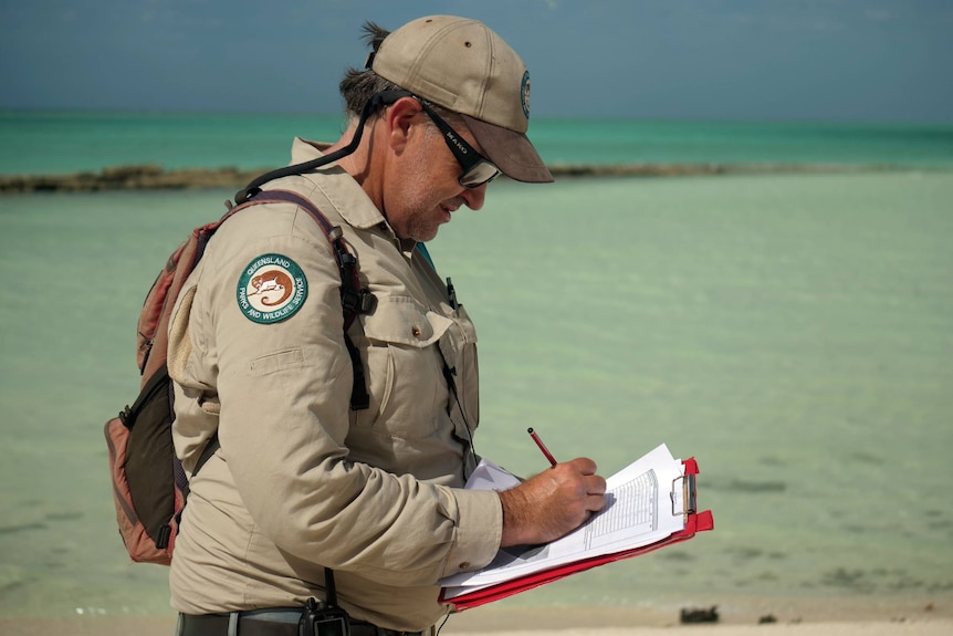 Damon wearing his khaki qpws uniform, writing on a clipboard, stunning clear water, sand behind.