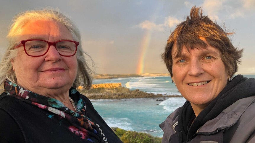 Two women stand near a bay with a rainbow behind them.