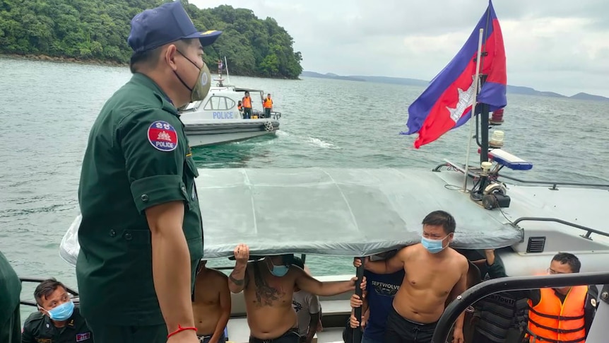 A small group of shirtless Chinese men wearing masks emerge from a small boat onto a dock as Cambodian police watch on.