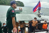 A small group of shirtless Chinese men wearing masks emerge from a small boat onto a dock as Cambodian police watch on.