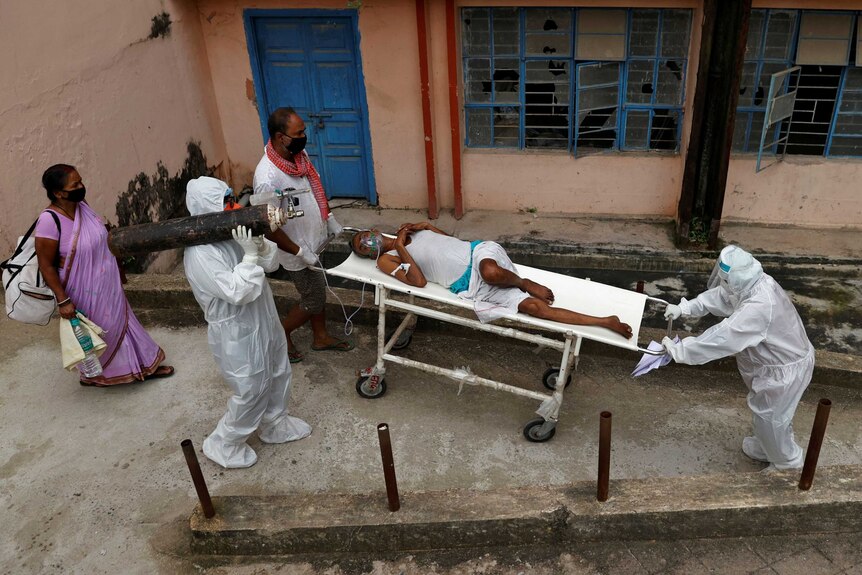 A man is wheeled on a stretcher as another in scrubs carries an oxygen tank while two men pull the stretcher