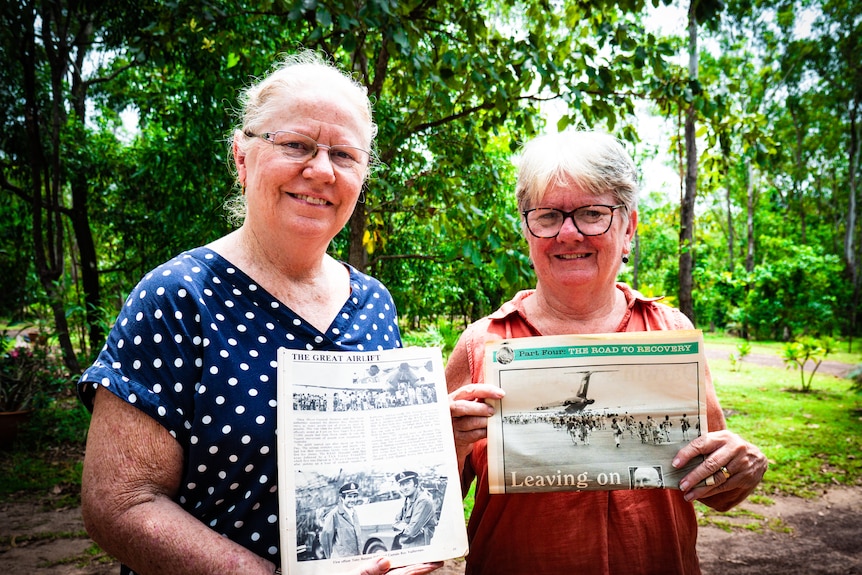 two women in a lush tropical garden holding newspaper clippings.