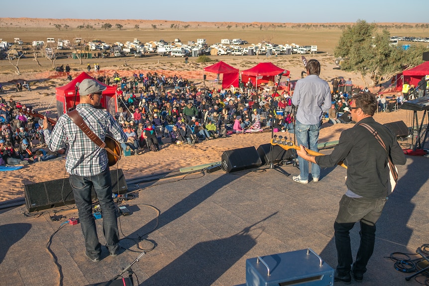 Three men play music for a small crowd in the desert.