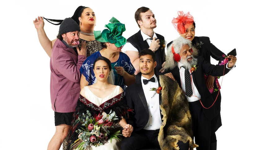As a bride and groom in formalwear pose for the camera, six other people pose for a selfie on a mobile phone behind them.