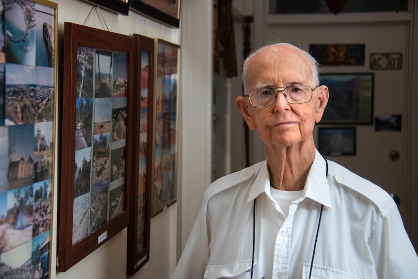 An elderly man looking at the camera, standing next to a wall full of framed photographs, wearing glasses and a white collared shirt