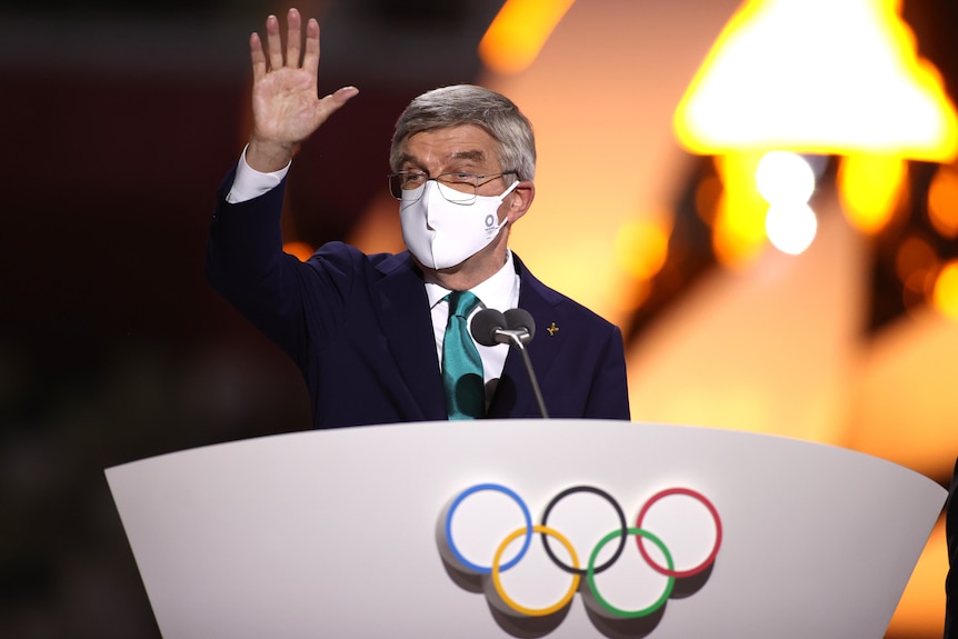 The IOC president stands at a lectern wearing a mask waving to athletes at the closing ceremony of the Tokyo Olympics.