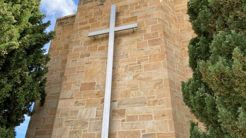 A large white cross hangs from the front of a church, either side stand two tall green trees