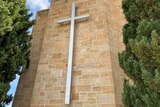 A large white cross hangs from the front of a church, either side stand two tall green trees