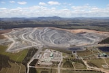 Aerial view of an open-cut coal mine. Treed hills and blue sky in distance.
