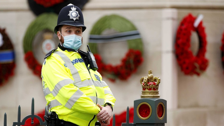 An English policeman in a face mask stands in front of a memorial covered in wreaths