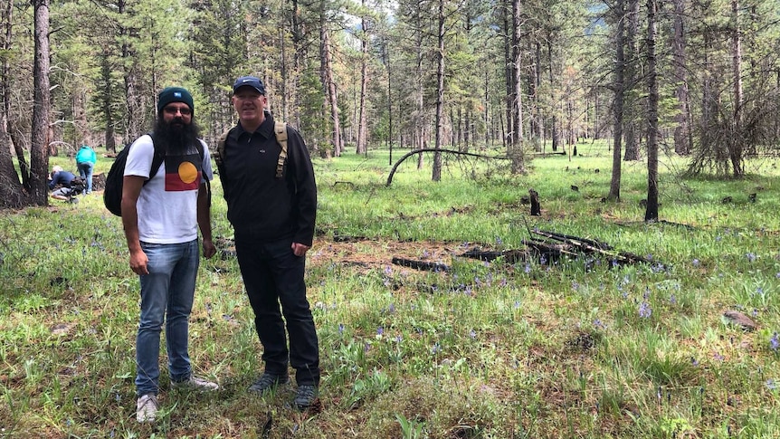 Two men stand in a forest clearing, smiling. In the background are pine trees and mountains