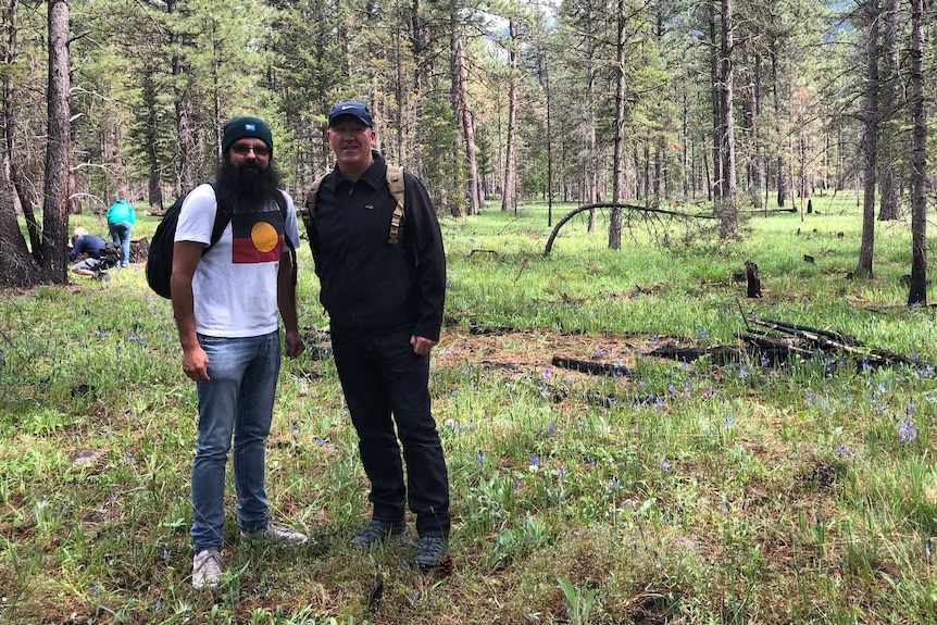 Two men stand in a forest clearing, smiling. In the background are pine trees and mountains