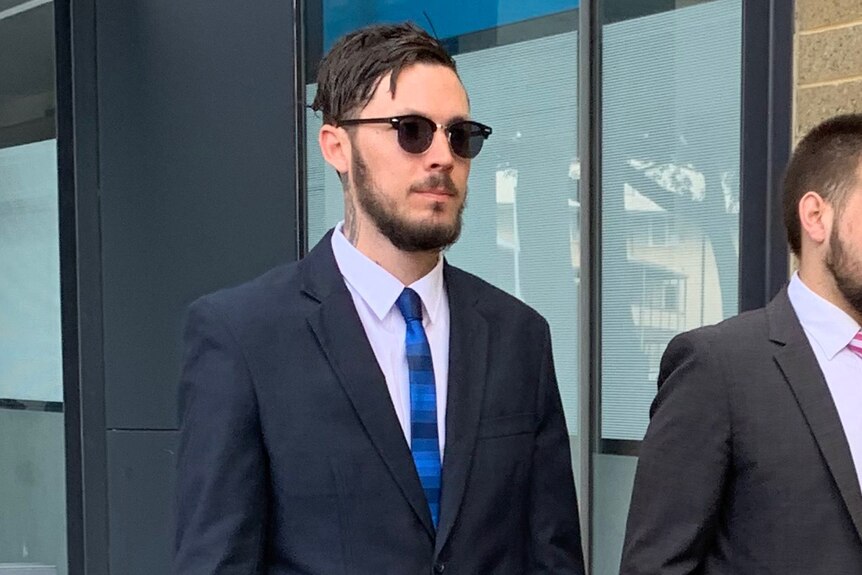 A mid shot of Mitchell Joseph Brindley walking outside a court building wearing a suit, tie and sunglasses.