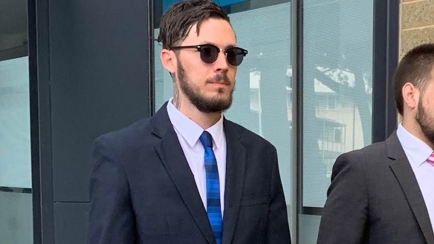 A mid shot of Mitchell Joseph Brindley walking outside a court building wearing a suit, tie and sunglasses.