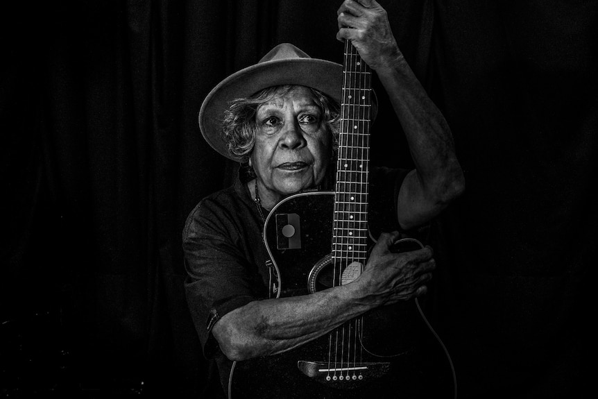 Black and white photograph of an older woman resting her face on an acoustic guitar staring out.