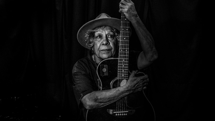 Black and white photograph of an older woman resting her face on an acoustic guitar staring out.