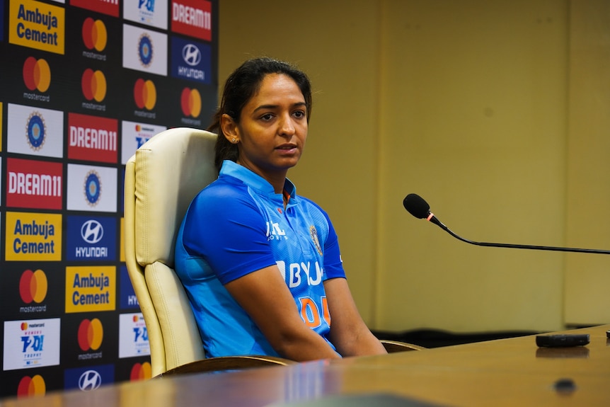 A woman wearing a blue cricket jersey sits on a chair behind a table answering questions.