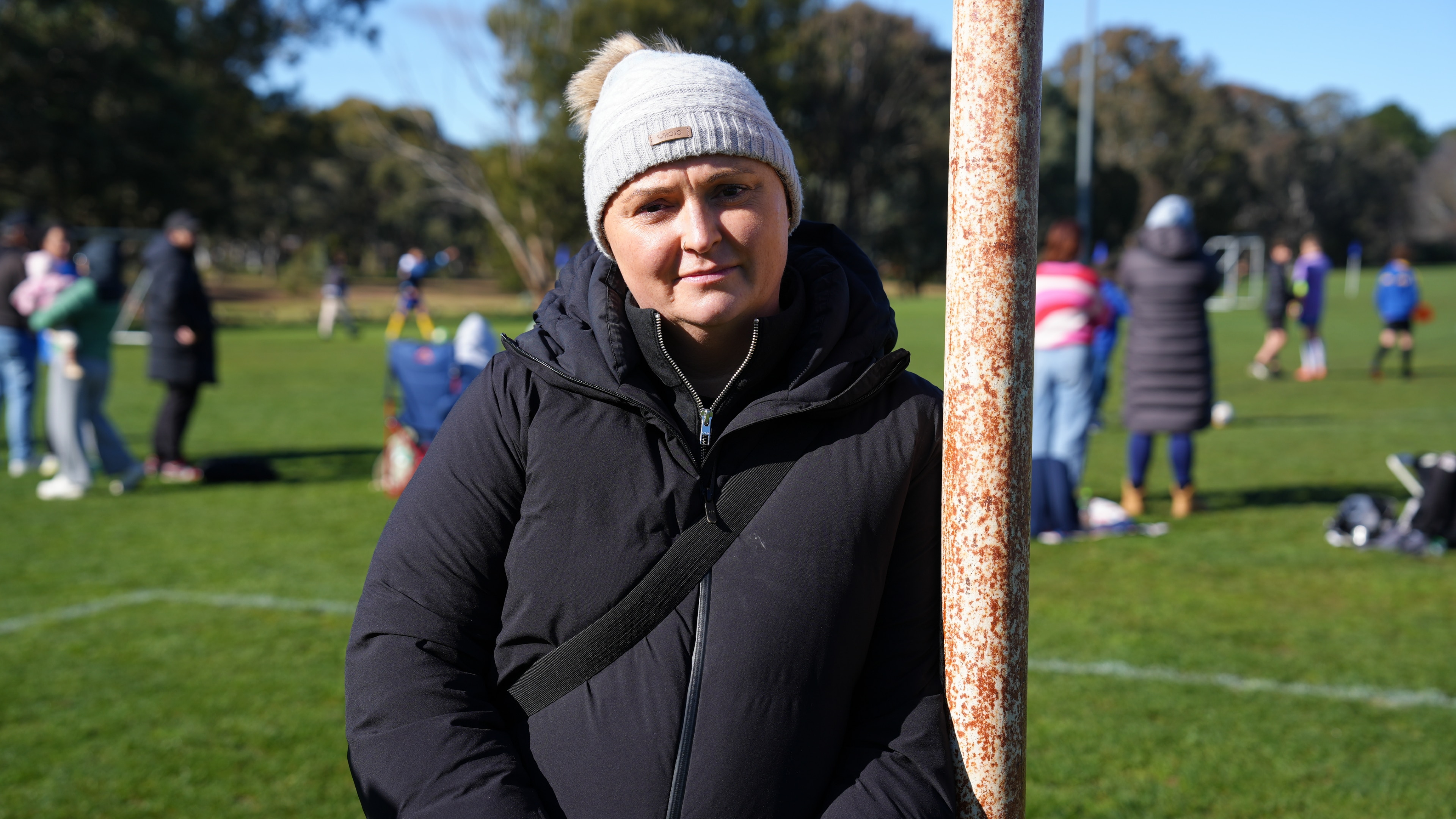 A woman wearing a beanie leans on a soccer field's goal post.