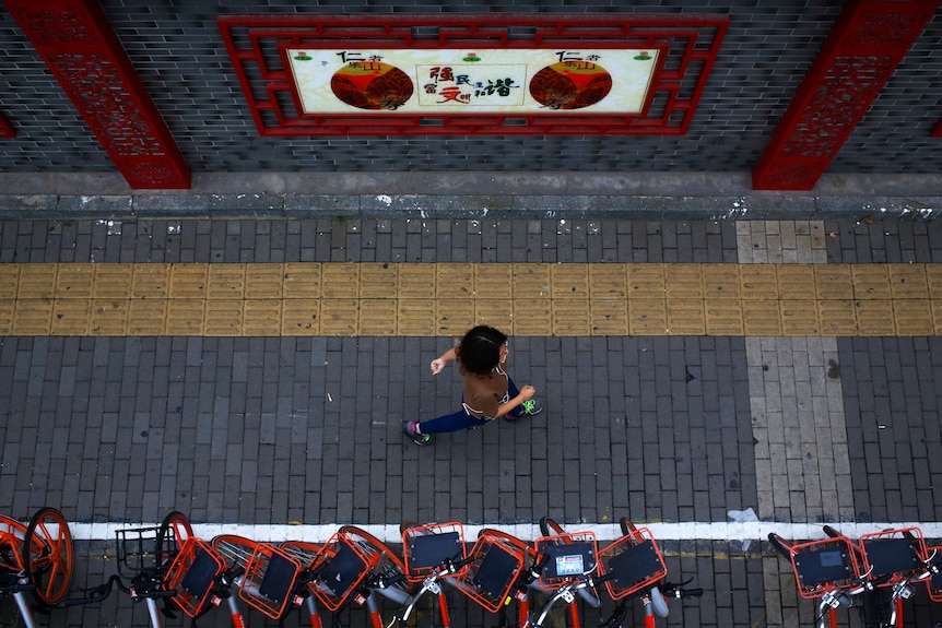 A woman walks by bicycles from a bike-sharing company parked along a wall displaying a government propaganda poster.