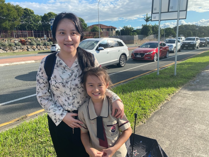 A woman stands on the sidewalk holding her smiling daughter by the shoulder. 