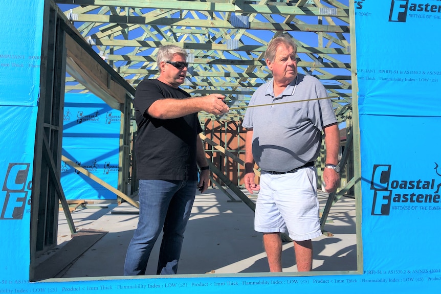 Two men stand in an incomplete house, one wearing jeans and a black T-shirt, the other in shorts and collared shirt 