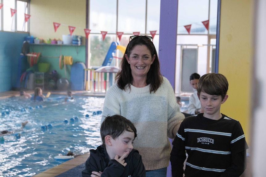 Ang Stewart stands in the door way with her two sons in front of her with an indoor swimming pool behind them.