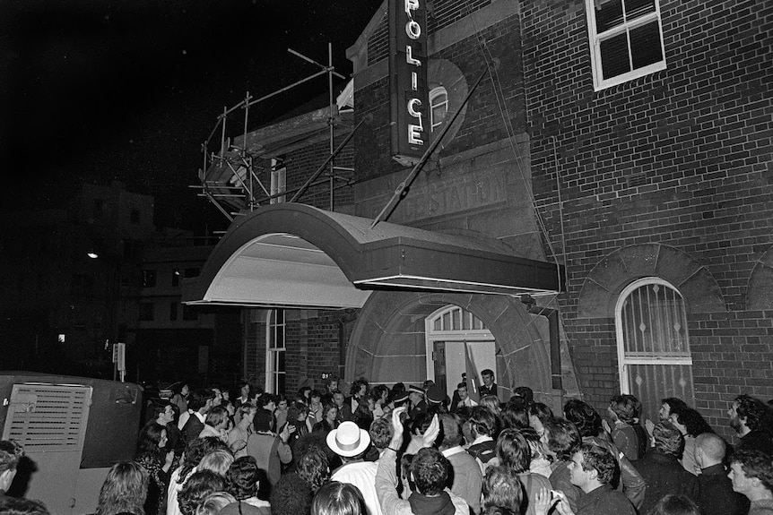 In a black-and-white photo, a large crowd stands at the door of the police station