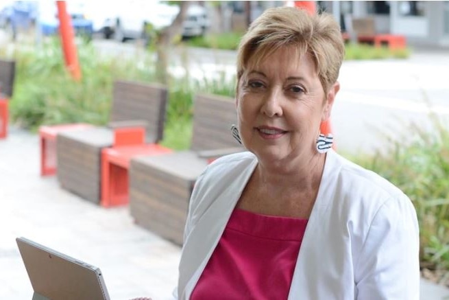 A woman with short hair, earrings, a white jacket and a pink shirt sits smiling.