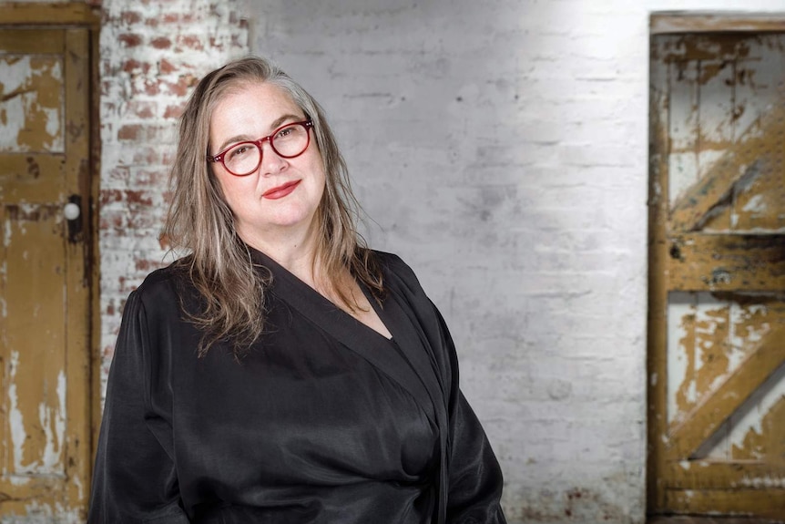 Woman with long greying hair standing with stripped back brick wall in background, head titled to side, red-framed glasses.