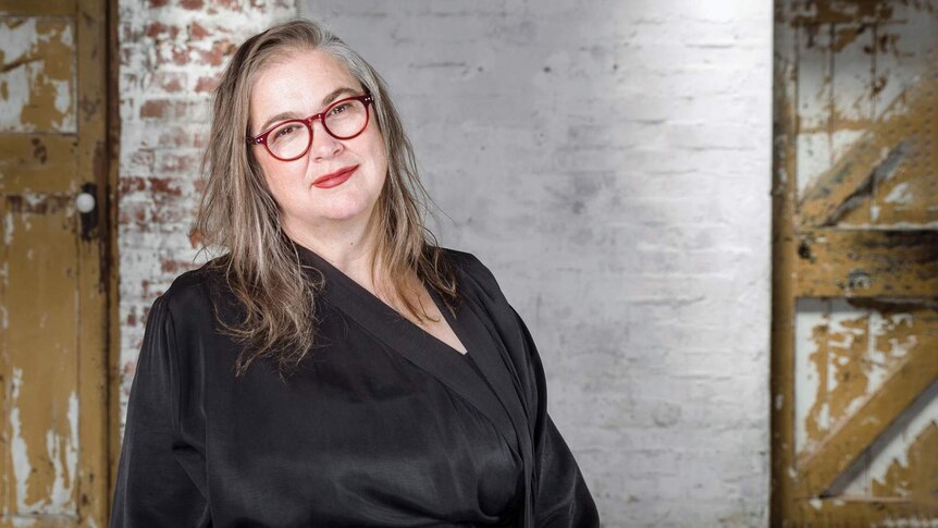 Woman with long greying hair standing with stripped back brick wall in background, head titled to side, red-framed glasses.