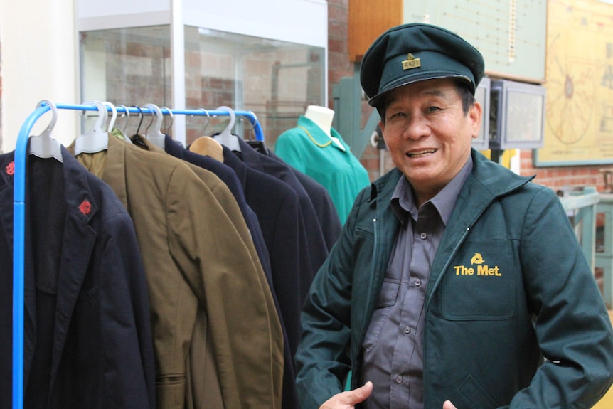 A smiling man wearing an old green tram conductor jacket and cap looks through other old uniforms at the Melbourne Tram Museum