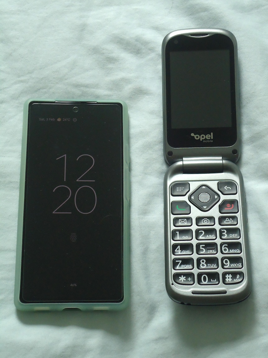 A close up of a smartphone resting on a bed, next to a flip phone which is open