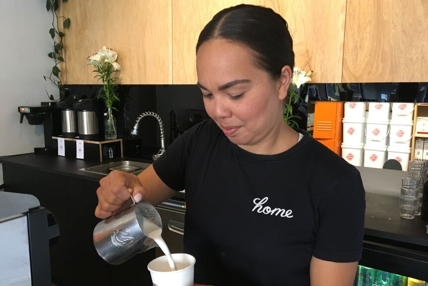 A woman in a black T-shirt in a cafe pours milk from a metal jug into a takeaway coffee cup.