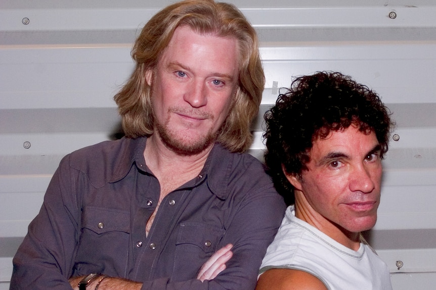 Daryl Hall smiles with his arms cross while a smiling John Oates leans against him