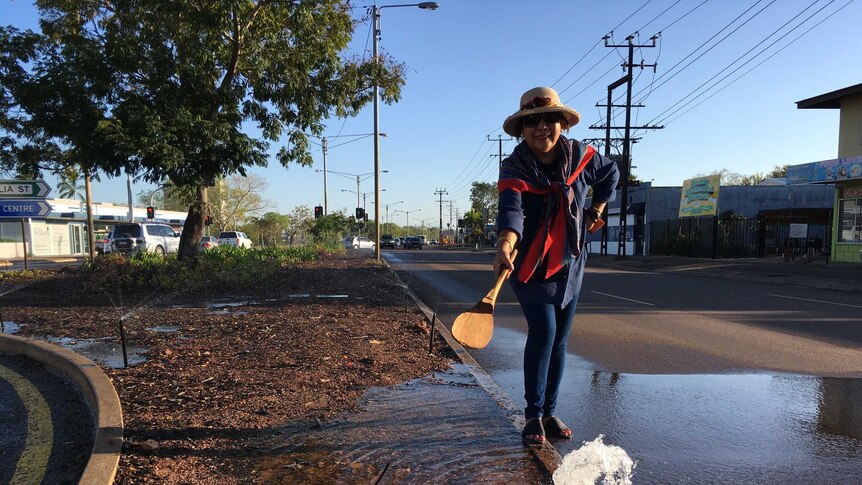 Laksa chef standing on a brown highway median strip with giant wooden spoon beside leaking irrigation