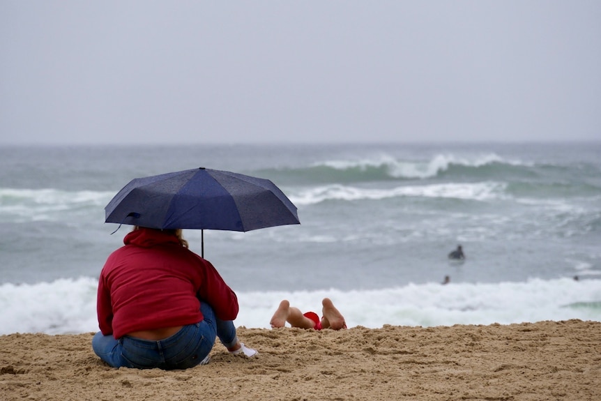 A woman sits at Burleigh Beach watching rough surf in the rain, while her child plays in sand.