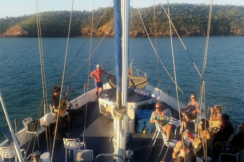 The deck of a large yacht showing people relaxing on chairs as a guitarist plays.