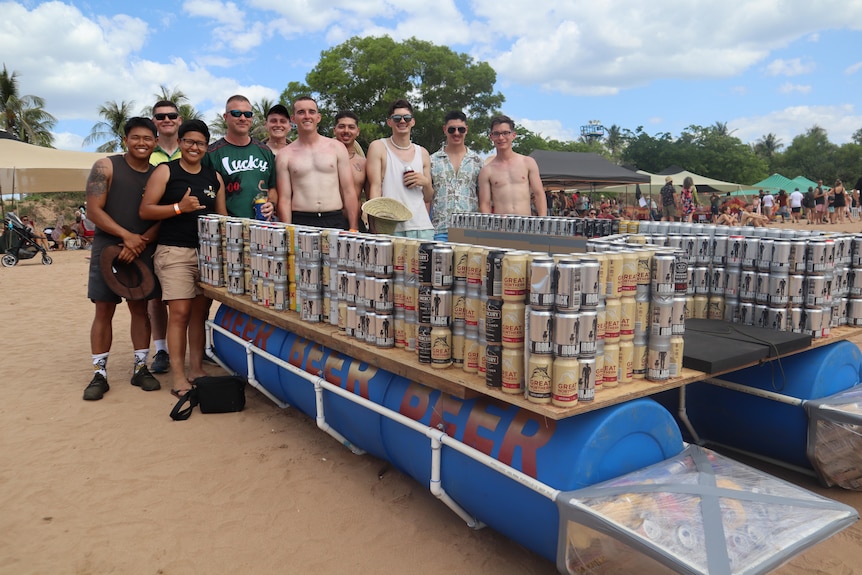 A group of men in their 20s, 30s and 40s standing behind a boat made out of beer cans, on a beach. 