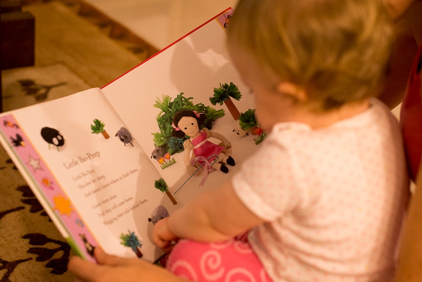 A toddler dressed in pyjamas sits on the floor pointing to a picture of a sheep on the page of a nursery rhyme book.