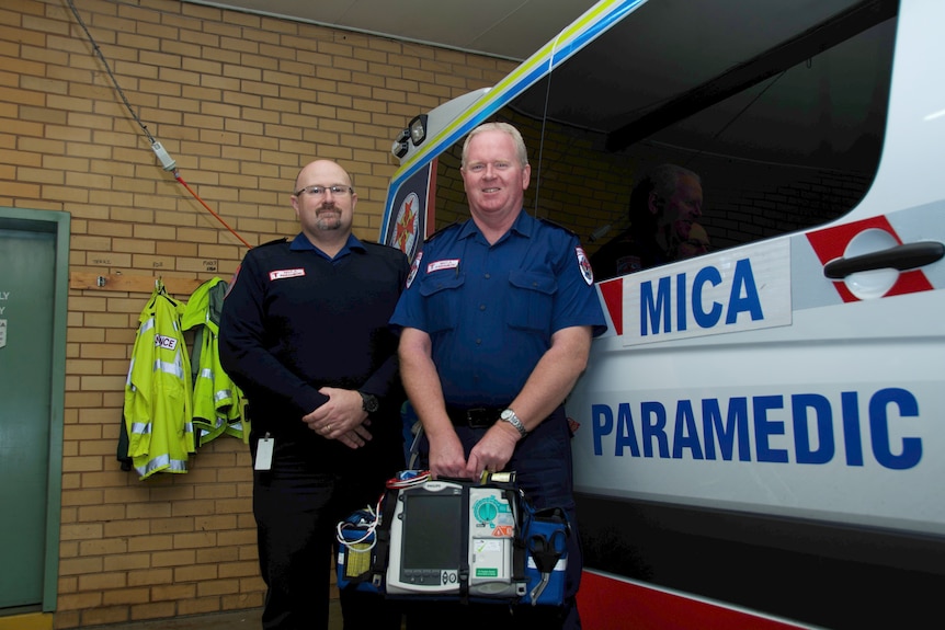 Two men in paramedics uniforms stand in front of an ambulance, smiling slightly.