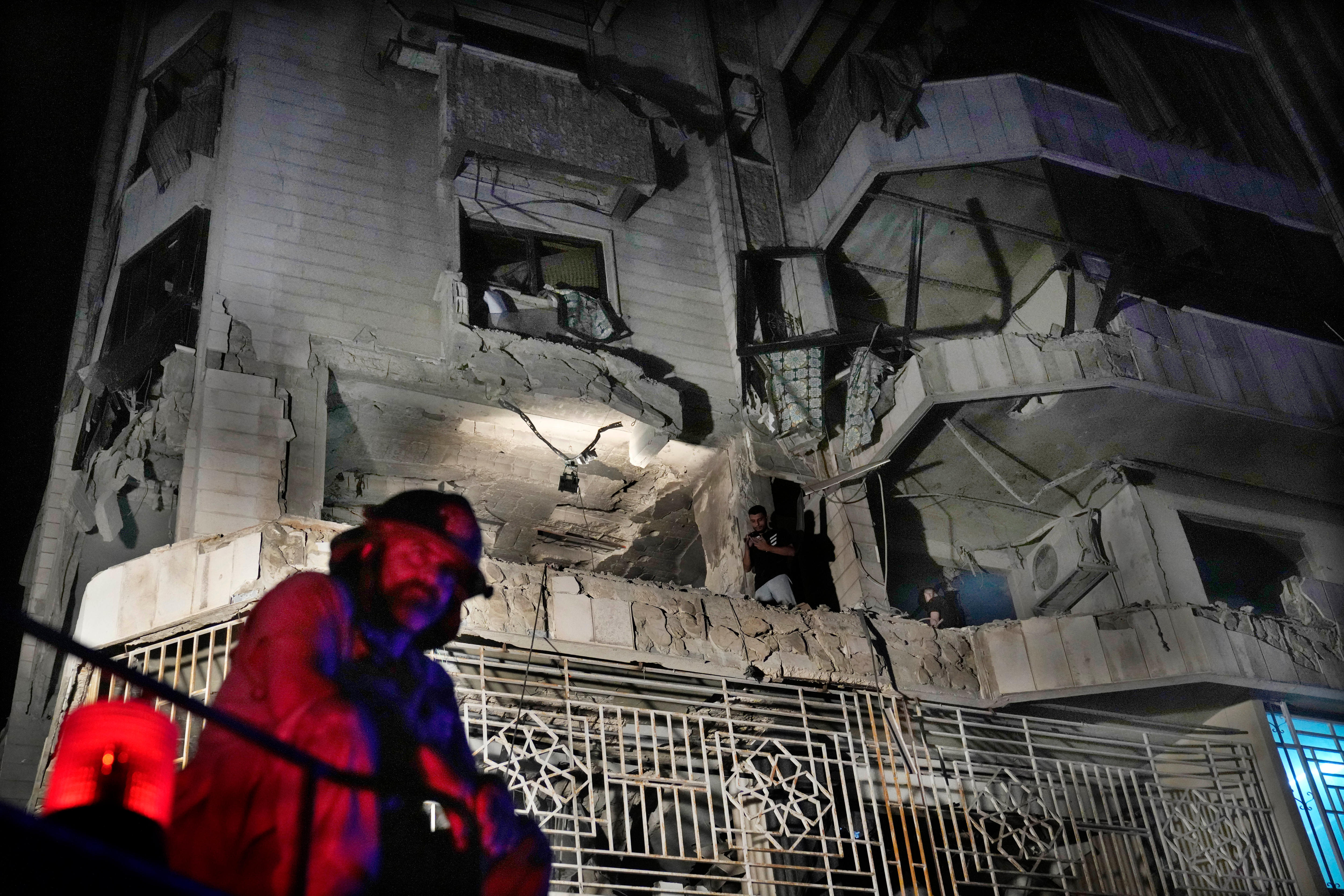 A bearded firefighter in shadow at night stands in front of a damaged building.