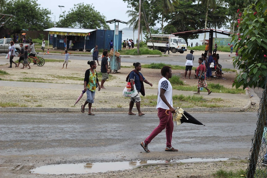 A man carrying an umbrella walks on the road, other people are walking in the background.