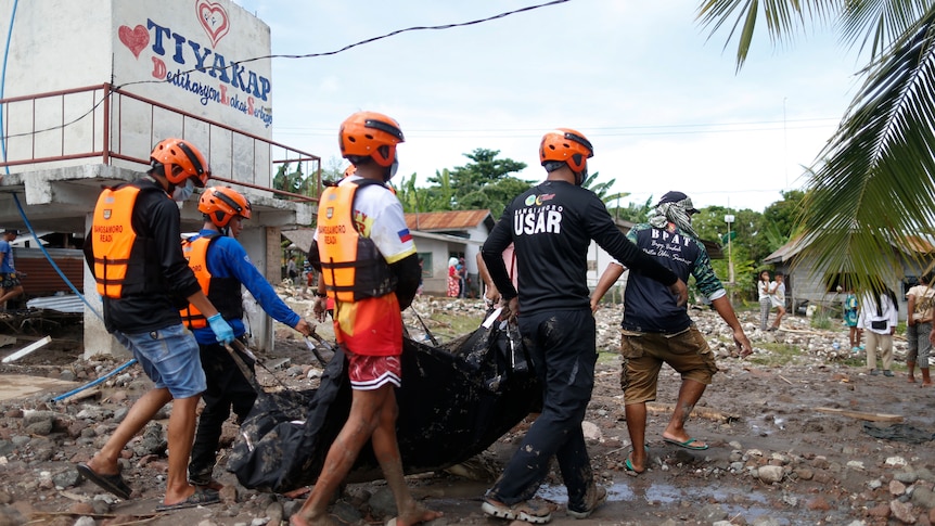 rescuers carry a black body bag through a street covered in rocks and mud