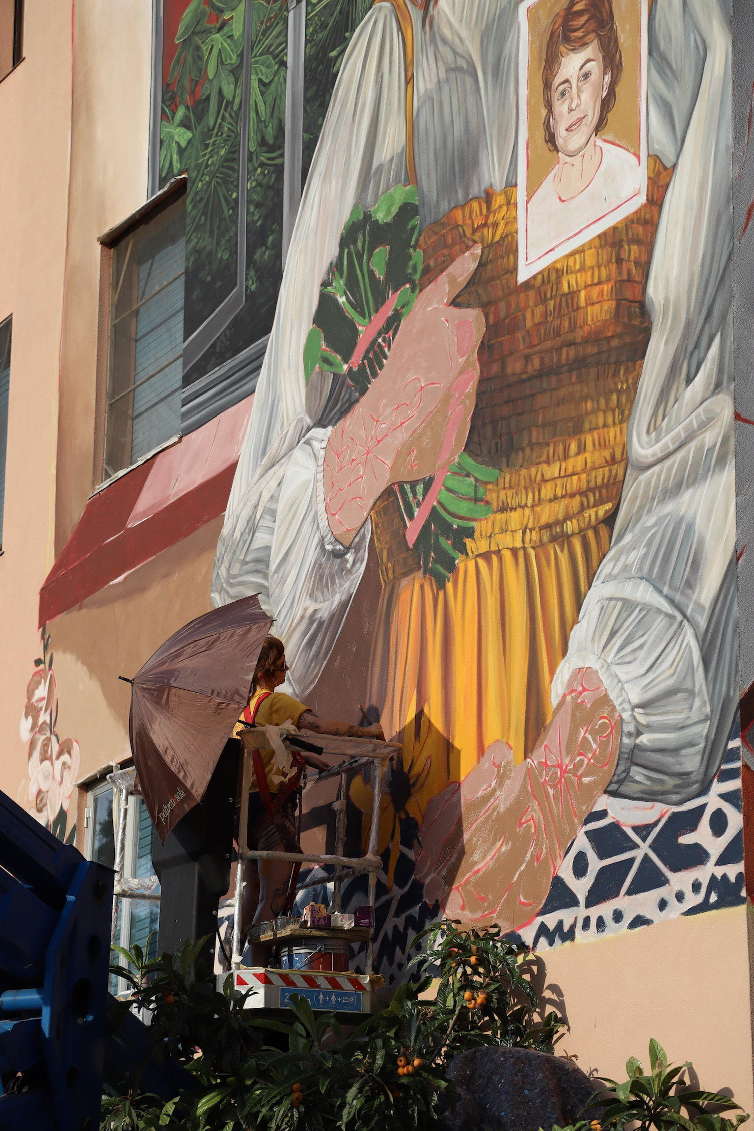 Artist stands on crane painting a mural on a large building. It is a sunny day and she is using an umbrella for protection