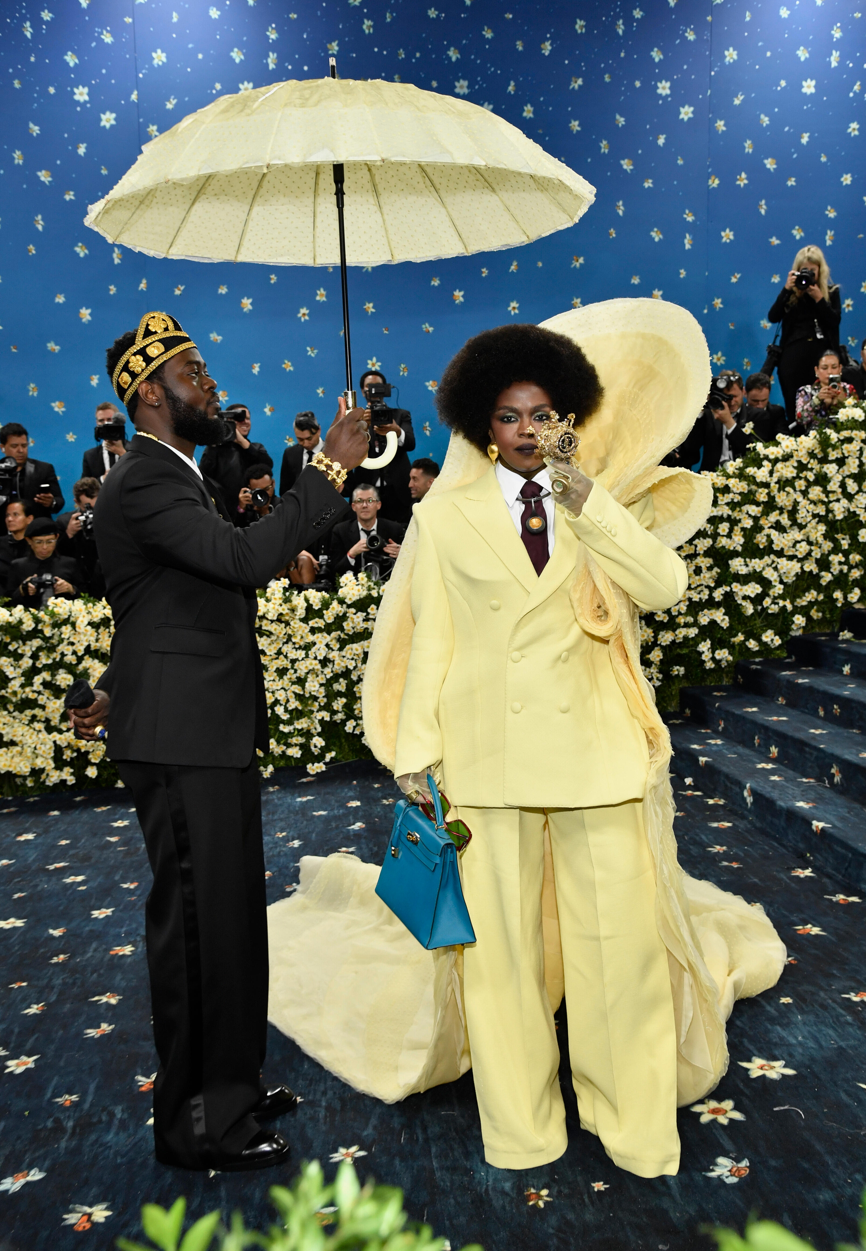 Lauryn Hill wearing a pale lemon yellow suit with a cape and an aide carrying a matching yellow umbrella