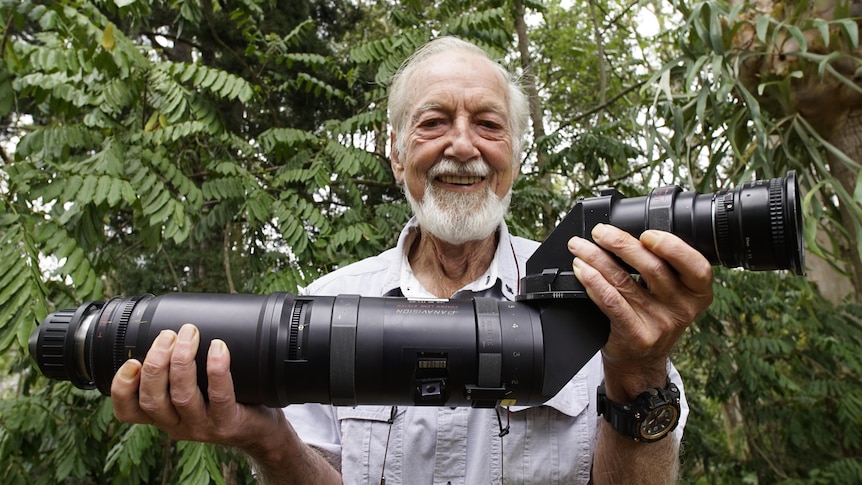 Man with grey hair smiles at the camera and holds a large camera lens