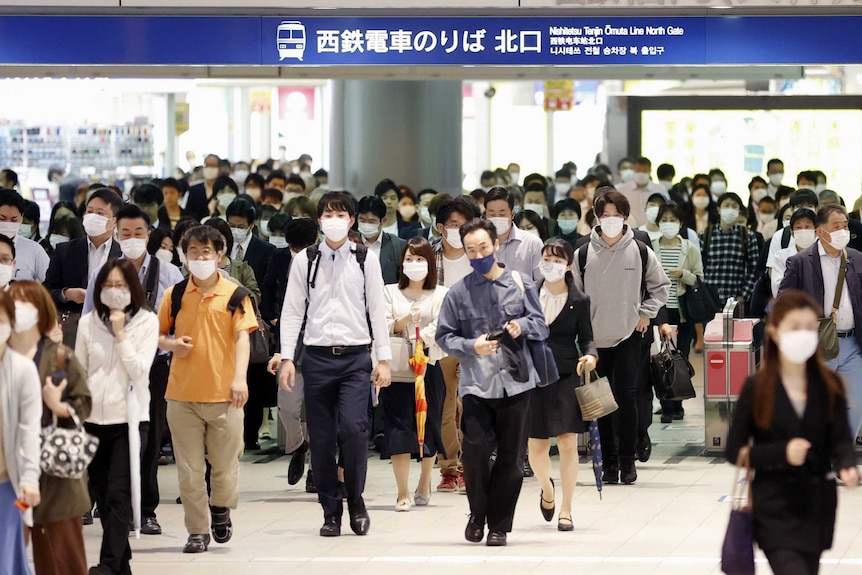 A crowd of people wearing face masks walk through a train station turnstile
