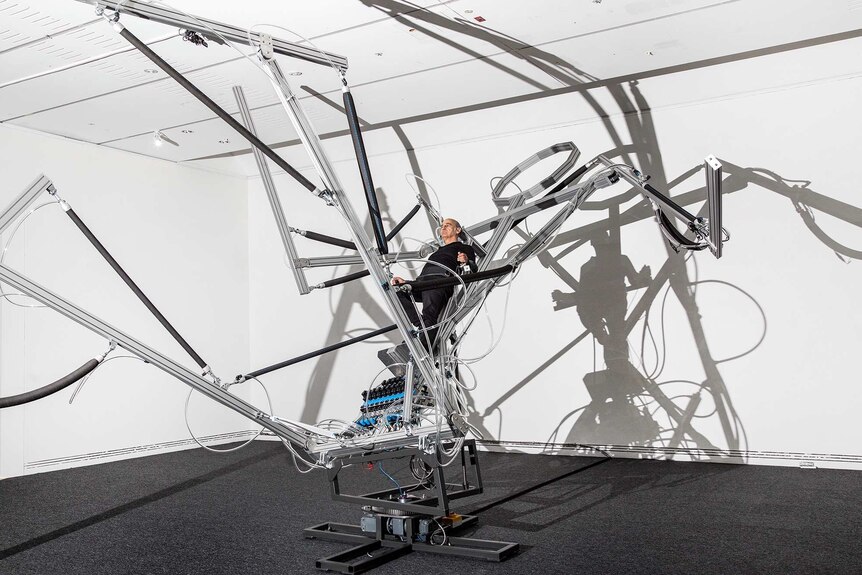 White gallery room with large robot-type machine in metal, and man sitting in centre of this, strapped in.