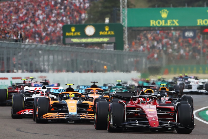 A Ferrari driver leads his car through a corner, with a stream of cars behind and packed grandstands in the background. 